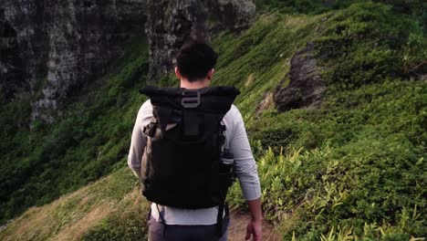 man hikes along tropical cliffside with large backpack, medium tracking shot