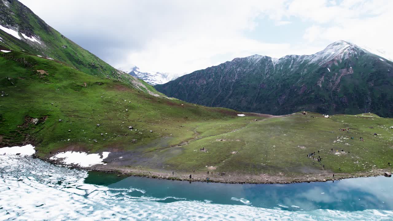 Frozen lake at the base of snow-capped mountains, bordered by lush green valleys