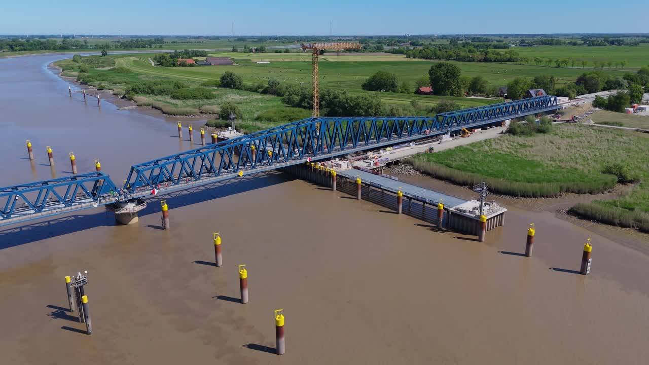 Aerial view of the new Friesenbrücke railway bridge over the Ems River in Germany. The swing-lift mechanism allows ship traffic to pass. Construction scene with piers, crane, and rural surroundings.