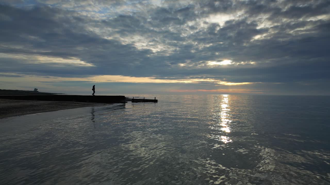 Milda vågor i slow motion med en manssiluett som går mot piruddens slut vid solnedgången på Fleetwood Beach i Lancashire, Storbritannien.