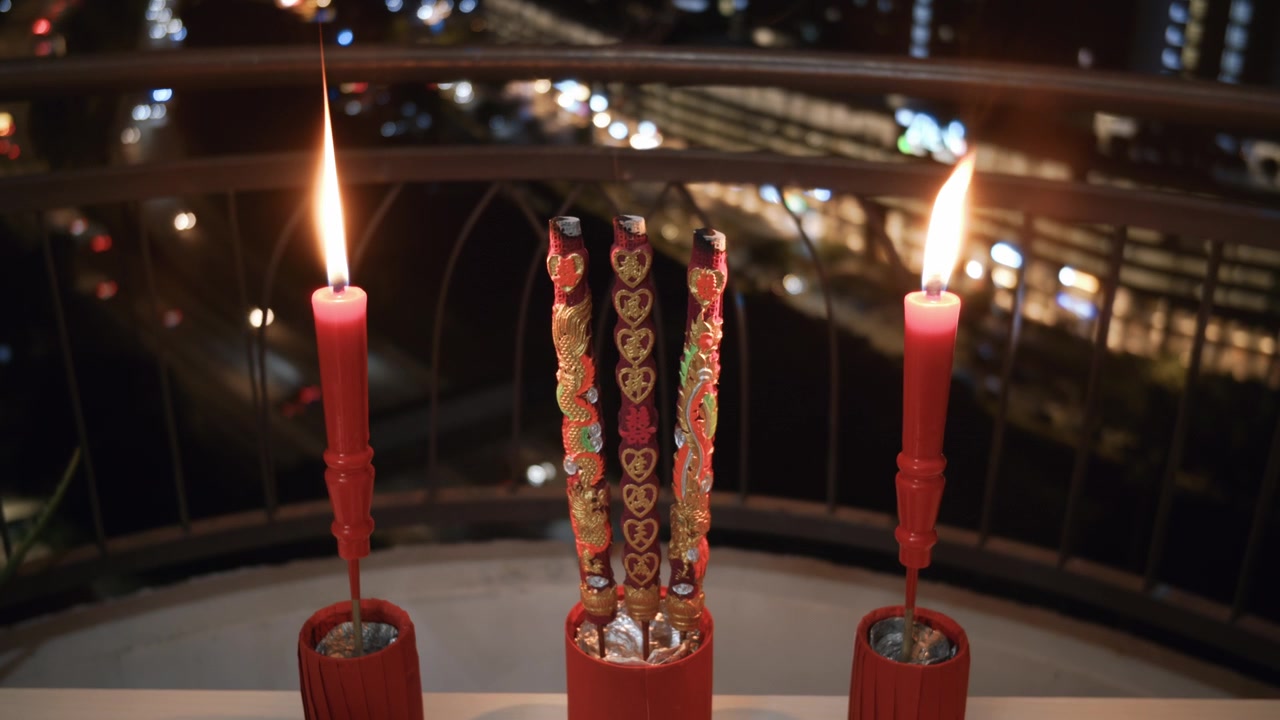 Tre bastoncini di incenso decorativi tradizionali e due candele rosse bruciano intensamente su un balcone con vista su uno skyline cittadino sfocato di notte