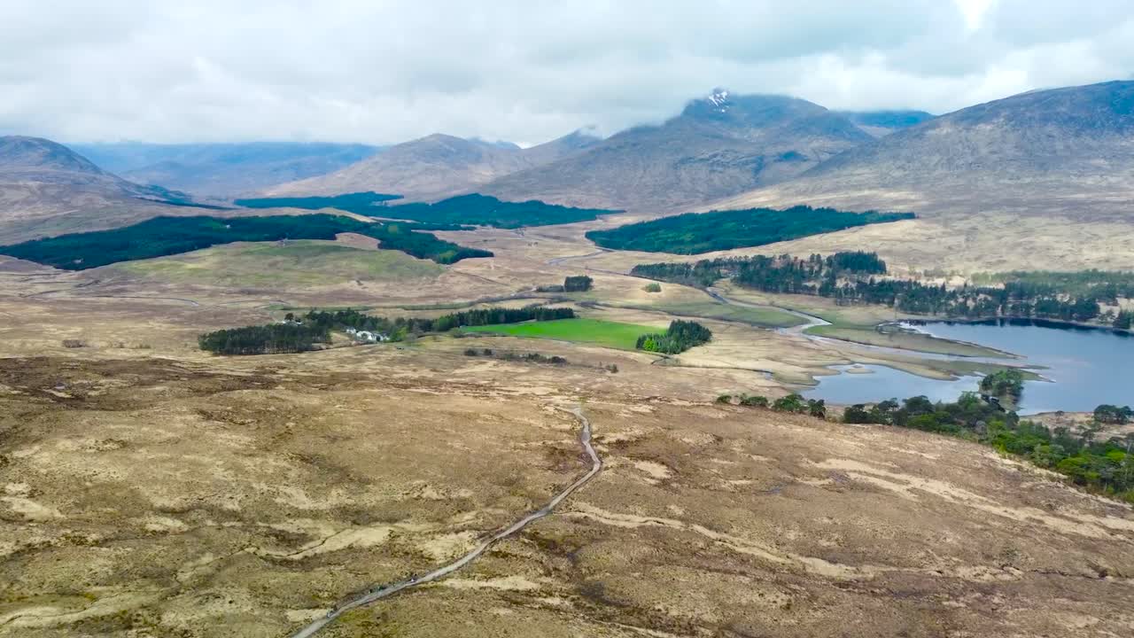 Aerial drone footage of gorgeous grassy brown scottish highlands landscape with a large blue calm water lake in the right hand side. Large hills and mountains visible in the background between clouds.