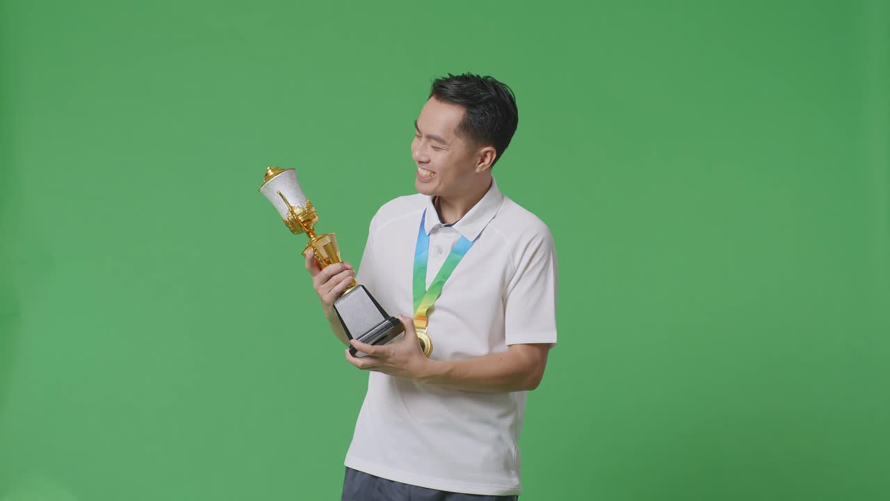 금메달을  아시아인 남자가 금 트로피를 키스하며 스튜디오의 녹색 스크린 배경에서 첫 번째 우승자로 자랑스러워합니다.