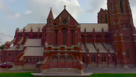 Una-Hermosa-Vista-Aérea-De-La-Iglesia-Antigua,-Un-Pájaro-Sentado-En-La-Cruz-De-La-Iglesia,-La-Iglesia-Con-Ladrillos-Rojos-Y-Dos-Autos-Estacionados-En-La-Esquina