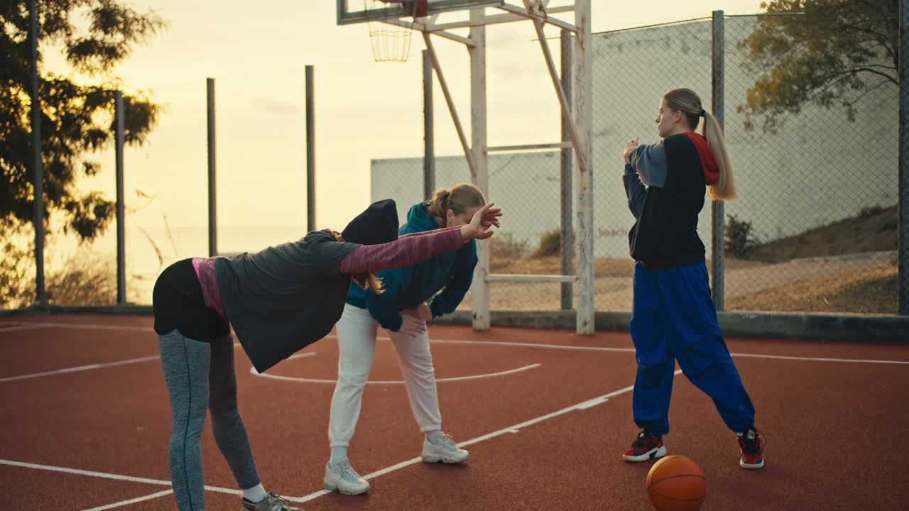 un trío de atletas y jugadores de baloncesto en ropa deportiva se están calentando en una cancha deportiva roja para jugar al baloncesto cerca de una pelota naranja temprano en la mañana