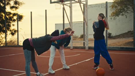 a trio of girls athletes and basketball players in sportswear are warming up on a red sports court for playing basketball near an orange ball early in the morning