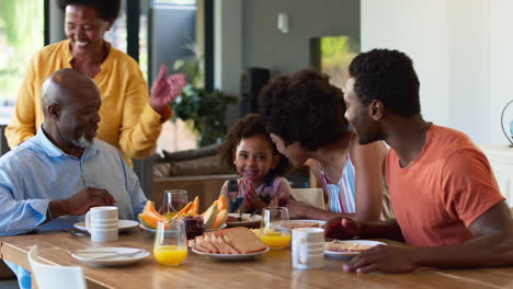 Family-Shot-With-Grandparents-Parents-And-Granddaughter-At-Breakfast-Around-Table-At-Home