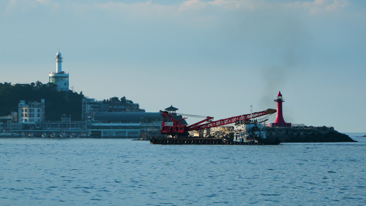 Grab Dredger Crane Ship Cruising In The Ocean Passing By On Red Lighthouse with Sokcho Lighthouse In The Distance