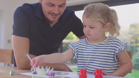 Father-and-young-daughter-having-fun-at-home-sitting-at-table-and-painting-decoration-together---shot-in-slow-motion