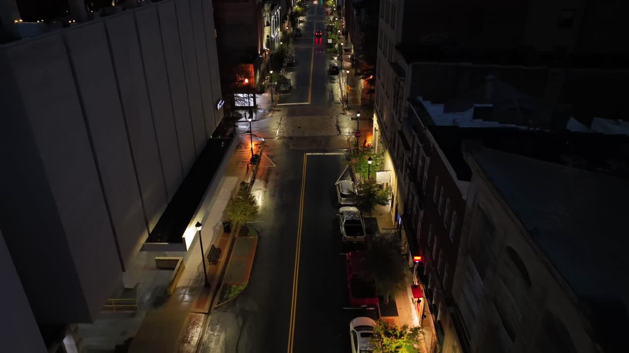 Aerial backwards shot of lighting street of american town. Glowing warm lighting streetlights with parking cars. Top down shot. Rainy day in autumn season.