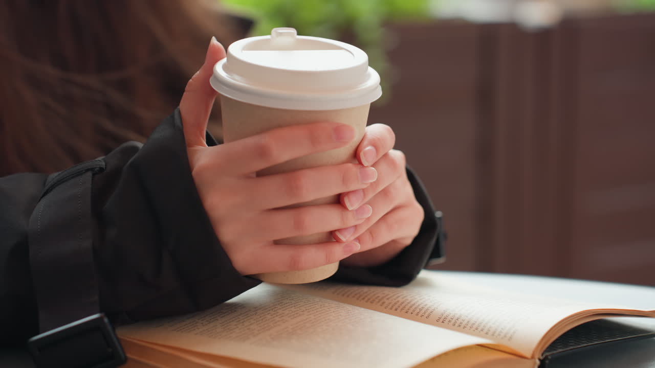 Donna che tiene in mano una tazza di caffè, libro aperto sulle gambe, luce mattutina soffusa. Primo piano delle mani di una donna che avvolgono una tazza da asporto, libro in brossura sul tavolo, capelli lunghi e giacca scura, vapore che sale, tranquillo patio urbano