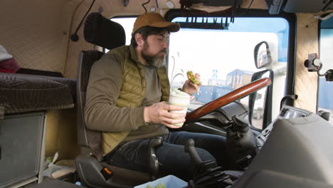 Side-View-Of-Worker-Sitting-In-A-Truck-In-A-Logistics-Park-While-Holding-A-Sandwitch-And-Drinking