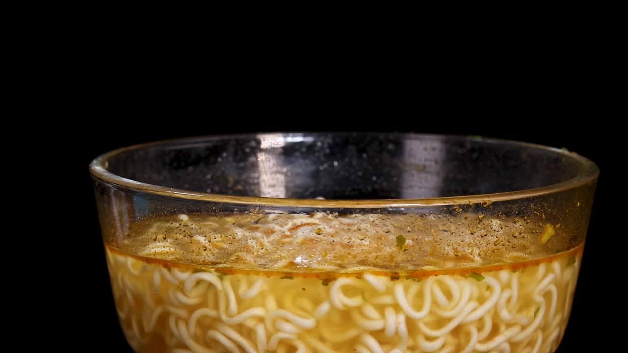 A glass bowl filled with instant noodle soup rotates smoothly against a solid black background, captured in even studio lighting with a steady camera angle