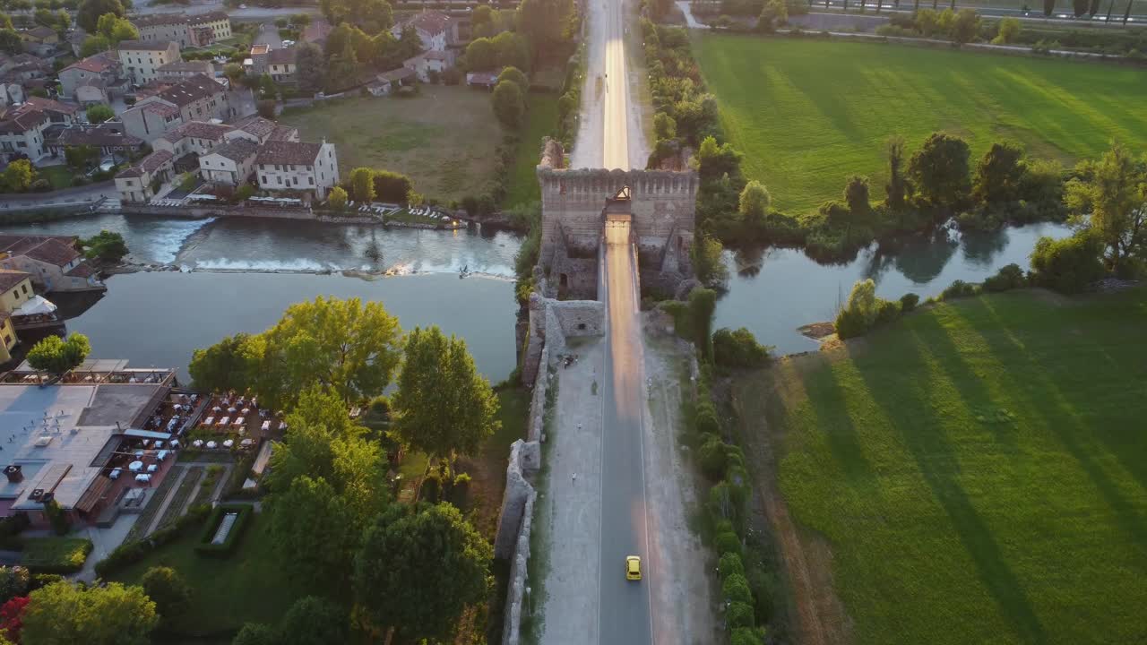 Golden hour aerial shot of Ponte Visconteo in Valeggio sul Mincio. The historic bridge stretches across the Mincio River, surrounded by green fields and medieval charm near Borghetto.