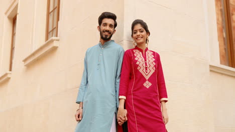 Portrait-of-happy-stylish-young-Hindu-man-and-woman-in-traditional-clothes-holding-hand-in-hand,-smiling-to-camera-at-street,-leaning-on-wall-of-building.-Beautiful-female-and-handsome-male.-India.