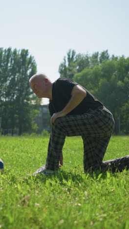 un anciano se arrodilla en una rodilla en un campo verde exuberante, centrándose en arreglar su zapato mientras una pelota de fútbol se encuentra cerca con árboles en el fondo