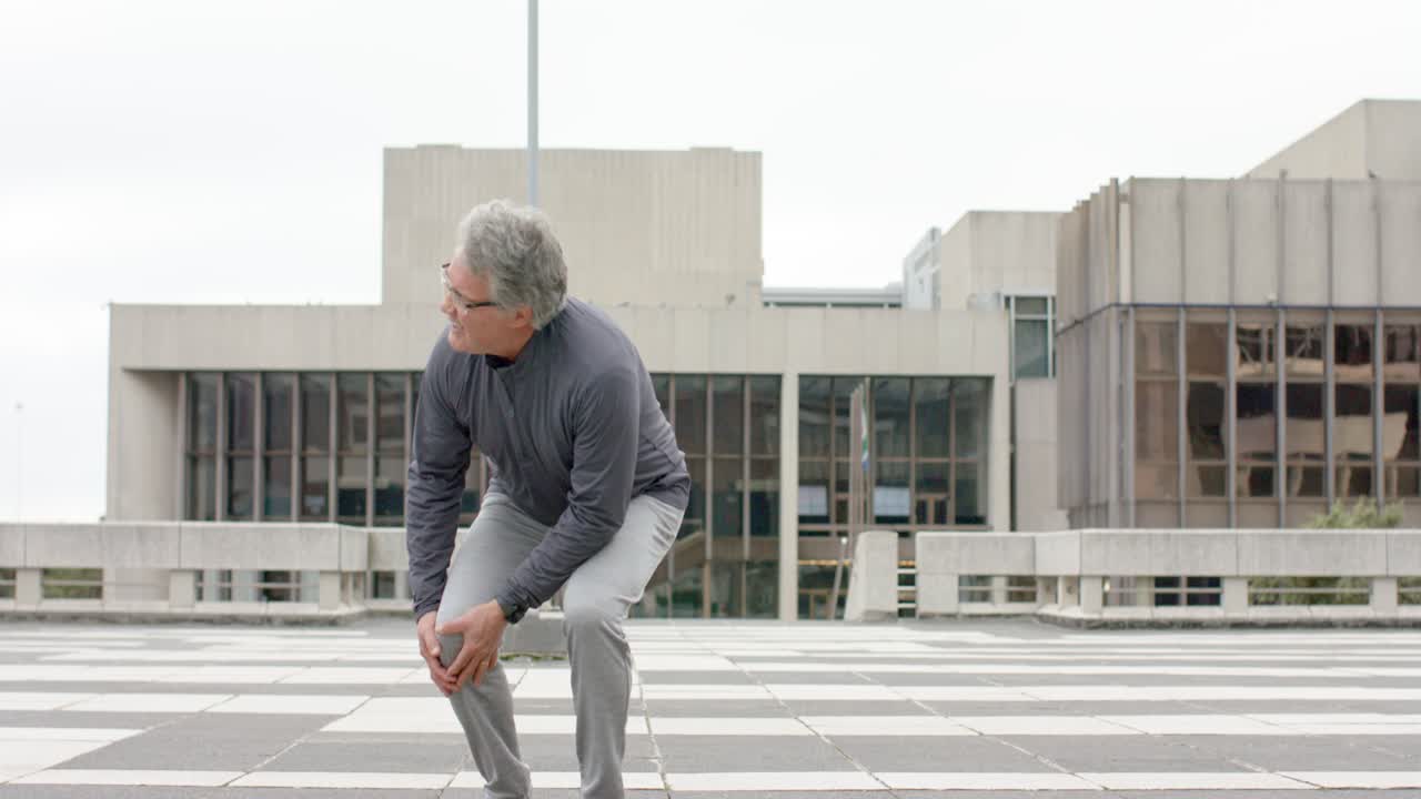 Jogging in sportswear across plaza, middle-aged male runner slowing and crouching after knee pain