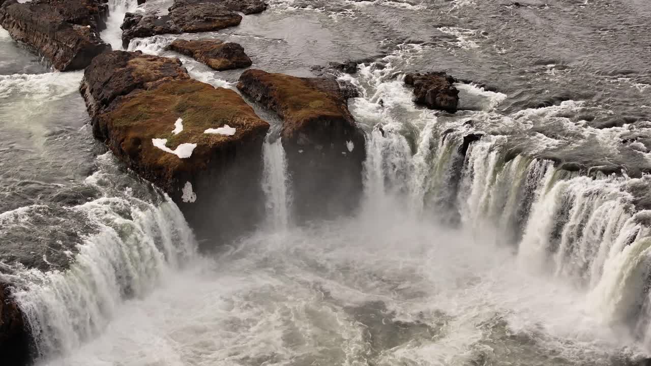 Close-up drone view of Goðafoss waterfall in Iceland, showing multiple powerful cascades plunging over rugged cliffs surrounded by patches of moss and snow in early spring.