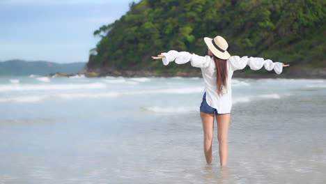 Rückseite-Einer-Sexy-Frau-Mit-Sommerhut,-Die-Sich-An-Einem-Sandstrand-Durch-Wellen-Ausbreitet-Und-Die-Hände-Hebt