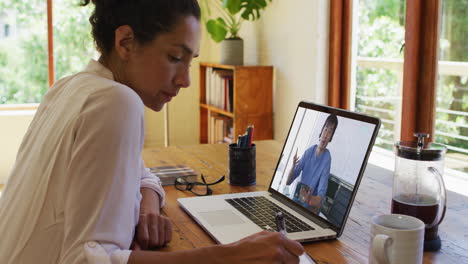 mujer de negocios biracial sentada en el escritorio usando una computadora portátil haciendo una llamada de video con un colega masculino