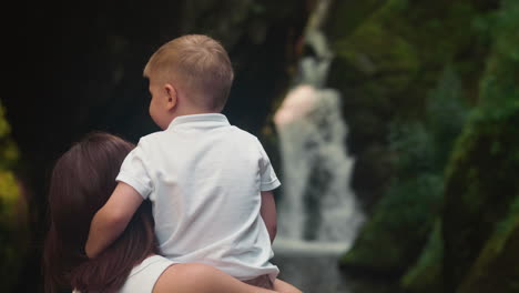 happy mother and child admire waterfall with clear water closeup backside view. woman holds boy in arms and child hugs mom neck tenderly slow motion