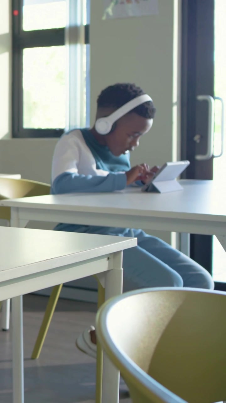 Vertical video: Tapping tablet, student with white headphones and blue top studying in classroom