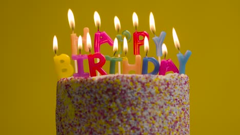 lit candles spelling happy birthday on cake covered with decorations against yellow studio background