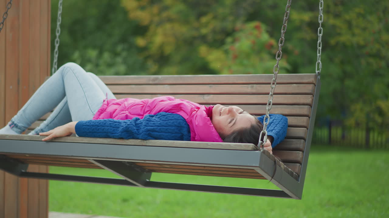 Donna caucasica sdraiata su un'altalena in un parco, che indossa un gilet rosa acceso e un maglione blu in maglia, jeans chiari e un berretto blu scuro, allunga il braccio verso il cielo, occhi chiusi, respiro rilassato, panca di legno sospesa.