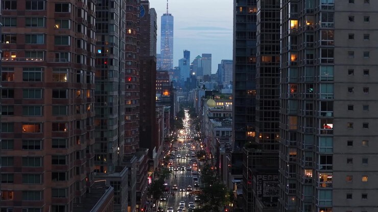 Luci serali illuminano un trafficato viale di Manhattan tra imponenti grattacieli residenziali, auto che scorrono sotto verso la sagoma distante del One World Trade Center. Ampia ripresa aerea con drone