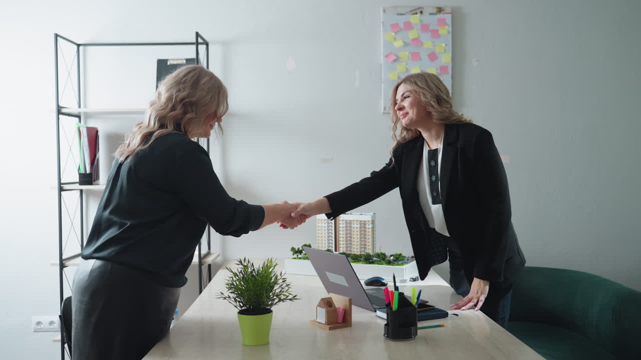 real estate agent seated in office welcomes guest with warm smile and gesture toward nearby chair, modern workspace setup with desk accessories, architectural model, laptop and green decor