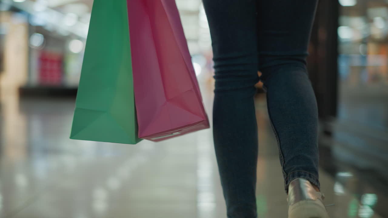 청바지와 캔버스 스니커를 입은 여성이 밝은 빛으로 조명된 쇼핑몰에서 는 동안 오른손에 민트와 분홍색 쇼핑 가방을 들고 있는 클로즈업 다리 시각