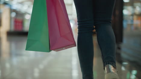 close-up leg view of a woman in jeans and canvas sneakers carrying a mint and pink shopping bag in her right hand while walking in a brightly lit shopping mall