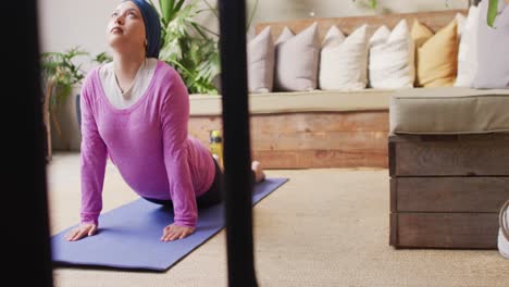 Video-Einer-Ruhigen-Birazialen-Frau-Im-Hijab-In-Yoga-Stretch-Pose-Auf-Dem-Boden-Des-Wohnzimmers-Zu-Hause