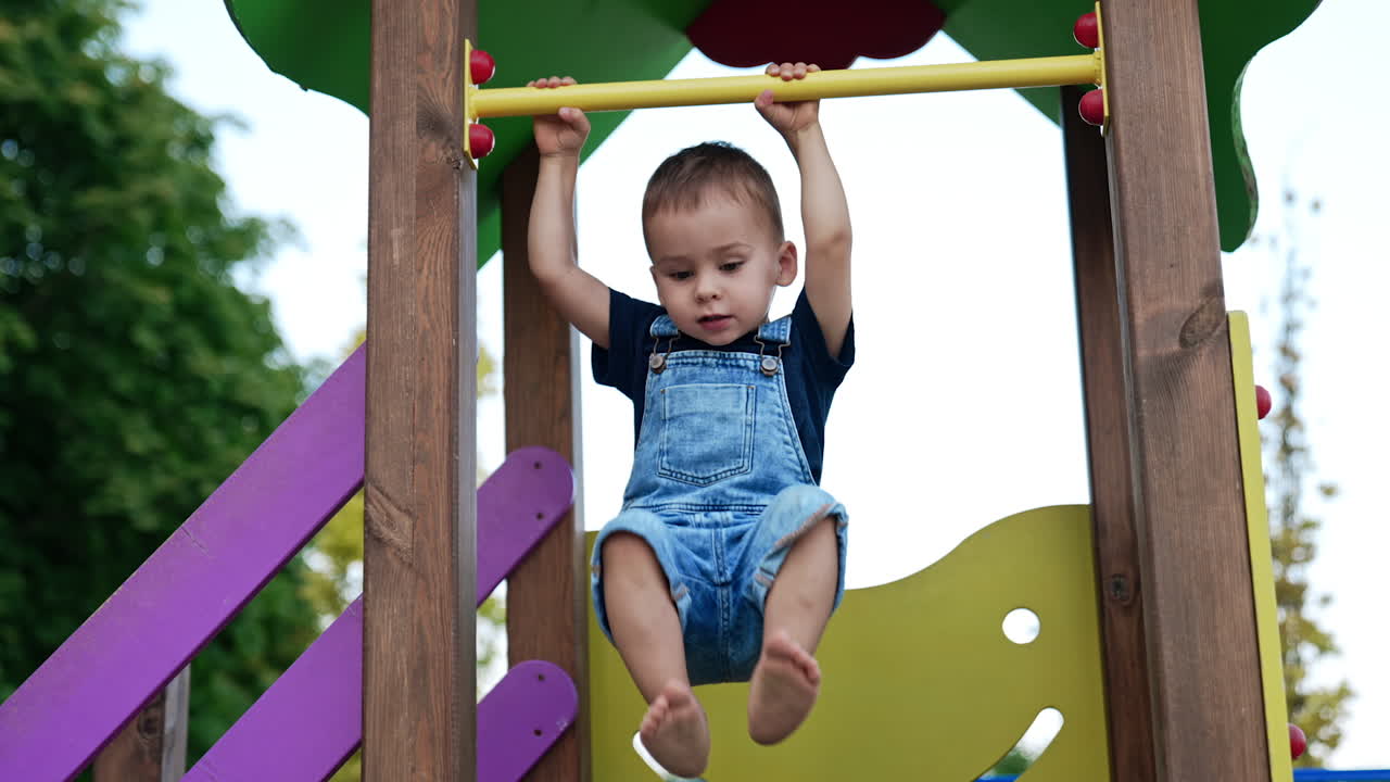 Happy smiling Caucasian toddler swinging on the horizontal bar. Active baby boy sitting on the slide at playground.