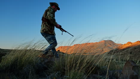 hunter wearing camo sits down in field with hunting rifle and takes aim