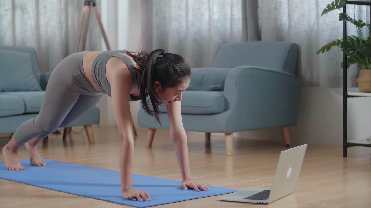 joven atleta asiática en ropa deportiva haciendo ejercicios de cardio rodillas altas y ejercicios de fortalecimiento del núcleo durante el entrenamiento en la alfombra en casa con entrenador en línea a través de una computadora portátil