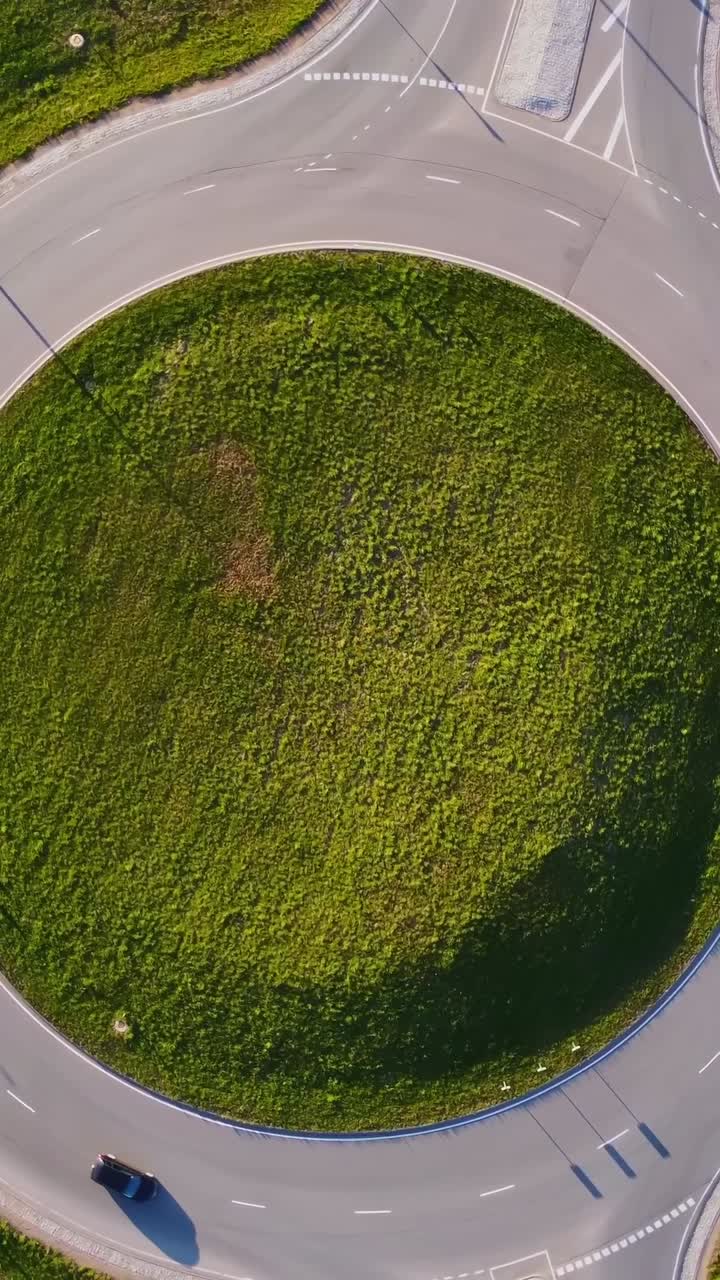 Cars driving on roundabout in Katlakalns residential area, surrounded by green grass and vegetation, on a sunny spring day in Riga, Latvia, top down vertical spining aerial view