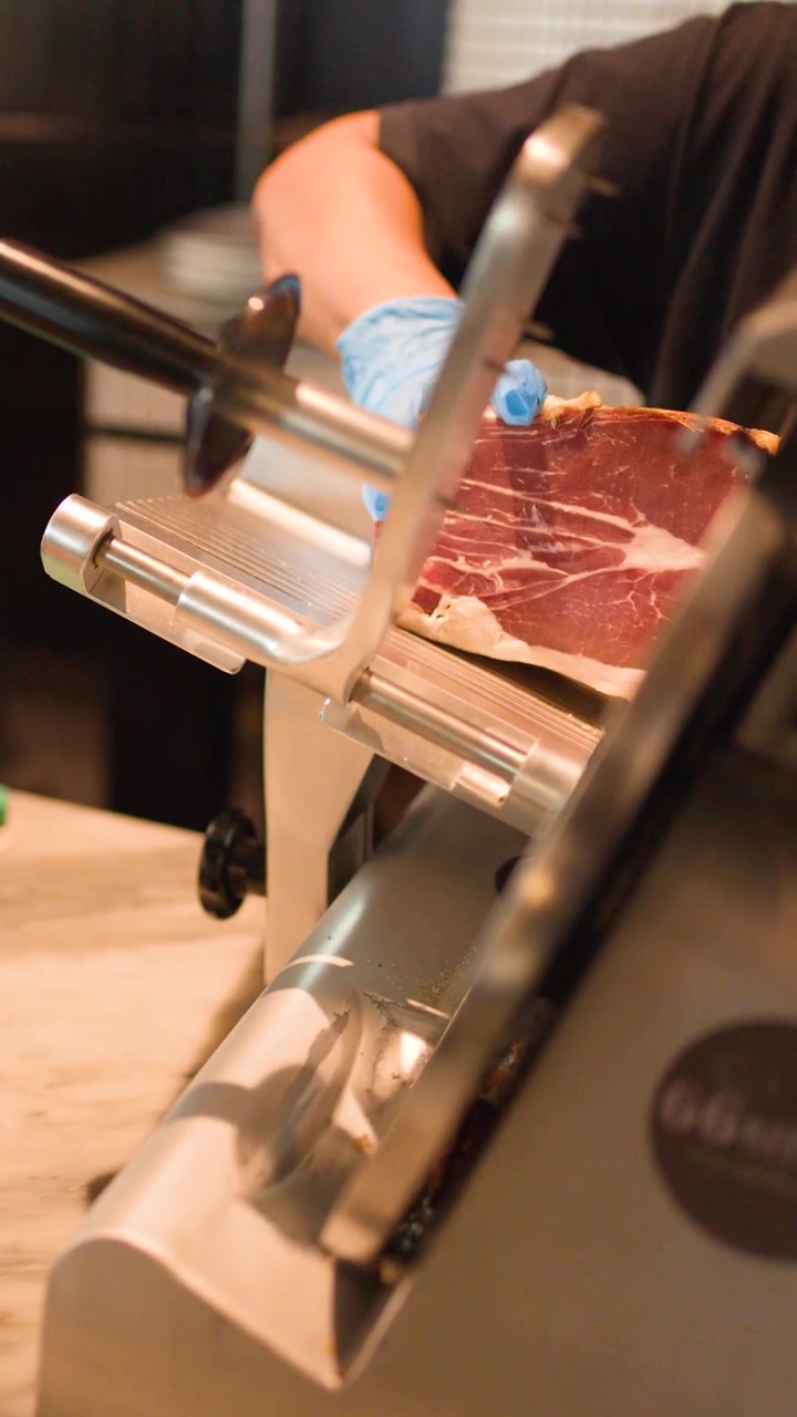 Primo piano di chef con guanti blu che usa un'affettatrice elettrica per fette sottili di prosciutto crudo.