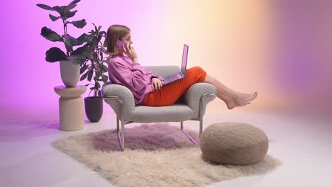 young woman in a modern pastel-lit room multitasks on a call while working remotely on her laptop, lounging comfortably in an armchair with plants and cozy décor around her