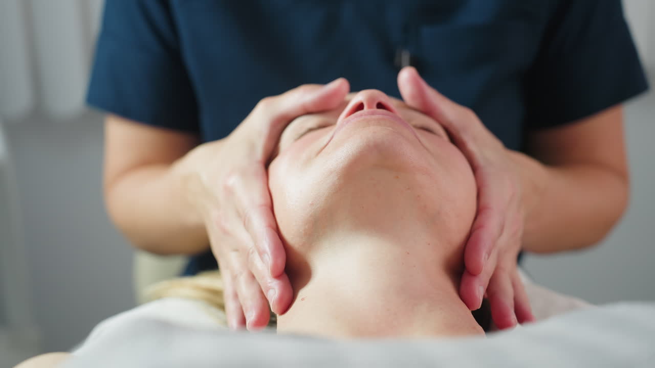 Treatment provider cradles customer jawline gently in bright spa room with white background during relaxing facial massage therapy session highlighting expert hands and serene wellness environment