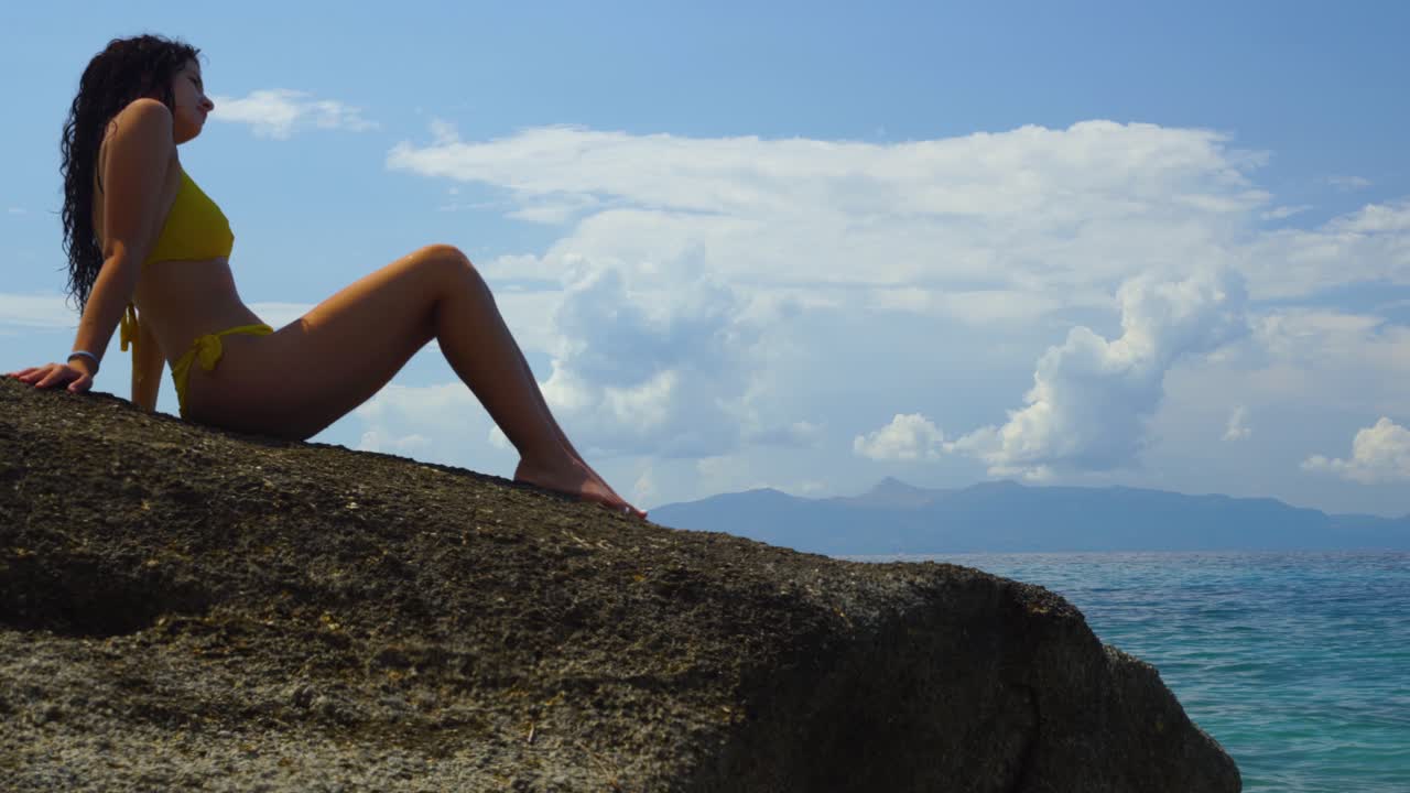 Ragazza in costume da bagno seduta su una scogliera, che osserva uno splendido paesaggio marino in un giorno di vacanza estiva.