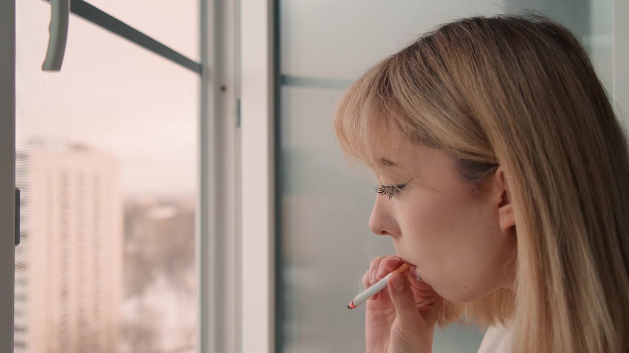 Donna riflessiva che fuma. Donna serena che guarda con una sigaretta. Donna contemplativa che riposa vicino alla finestra con una sigaretta accesa. Donna pacifica in pensieri silenziosi che fuma mentre la città si risveglia all'alba.