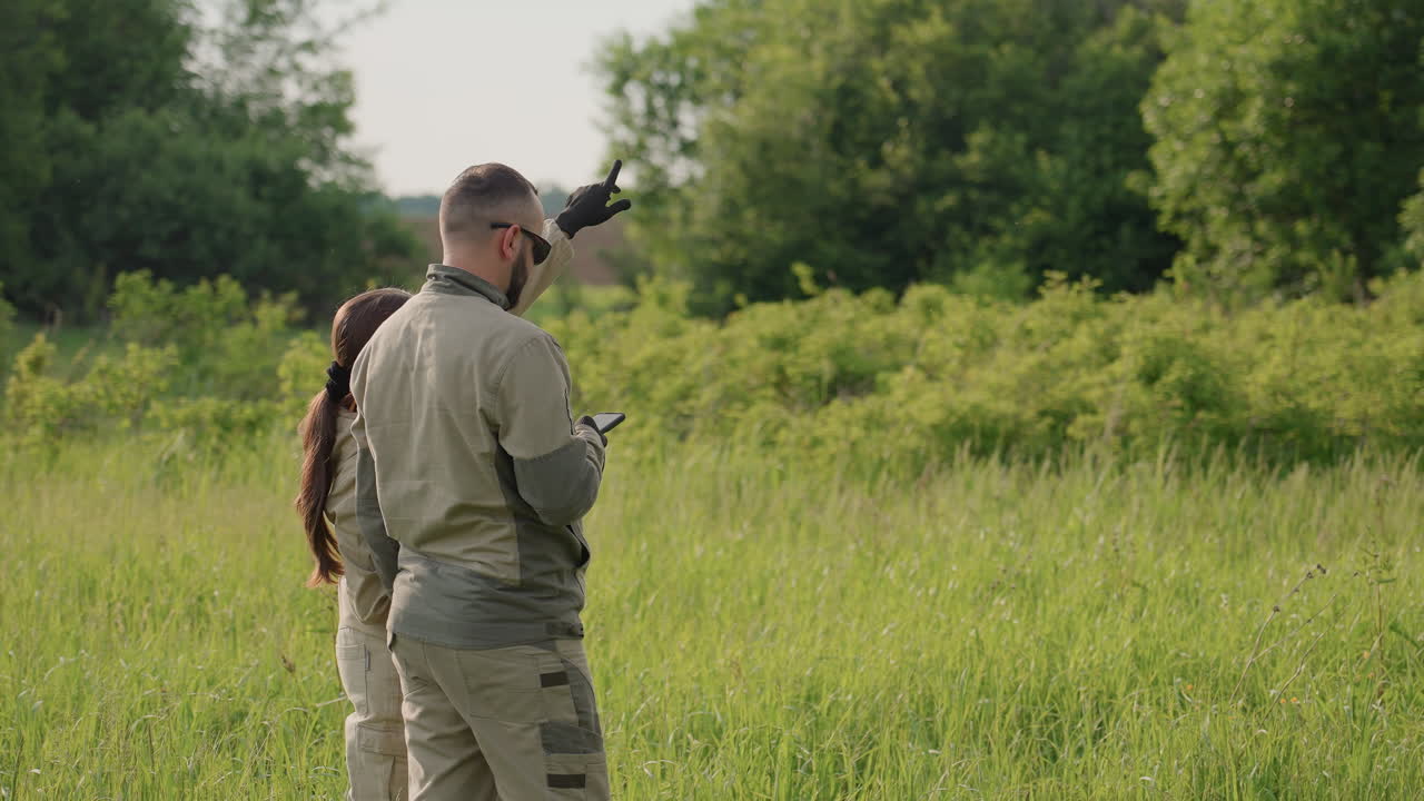 Researchers stand in tall grassy field holding device while scanning distant forest edge observing vegetation growth under bright summer daylight for scientific survey and data collection