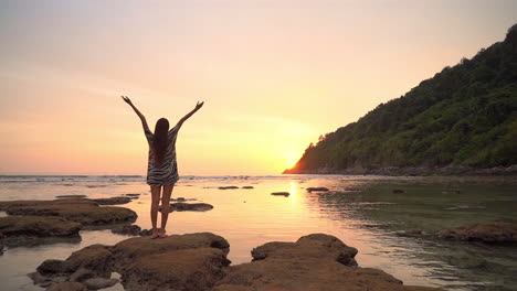 asian woman standing on rock by the sea water and raising hands up when the sun going down behind the mountain