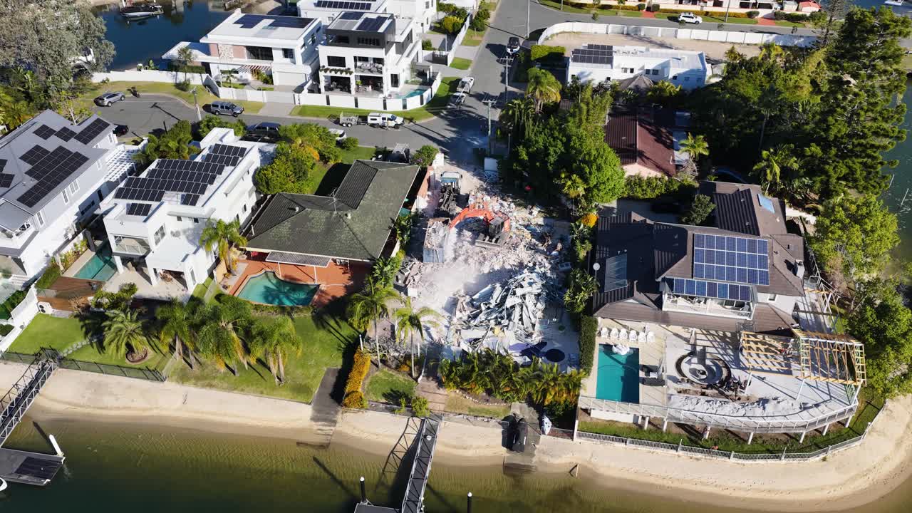 Drone footage captures a demolition machine tearing down a waterfront house in a sunny residential Gold Coast neighborhood, with rubble and machinery visible