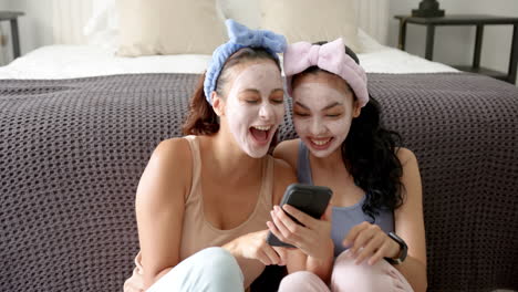 Two-young-biracial-female-friends-are-laughing-at-a-smartphone-with-facial-masks-and-headbands