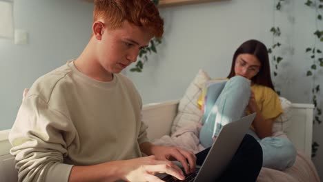 dos adolescentes caucásicos sentados en la cama y aprendiendo de libros y computadoras portátiles en silencio