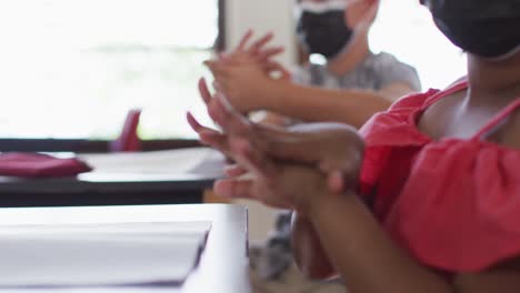 Diverse-schoolchildren-wearing-face-masks-disinfecting-hands-while-sitting-in-classroom