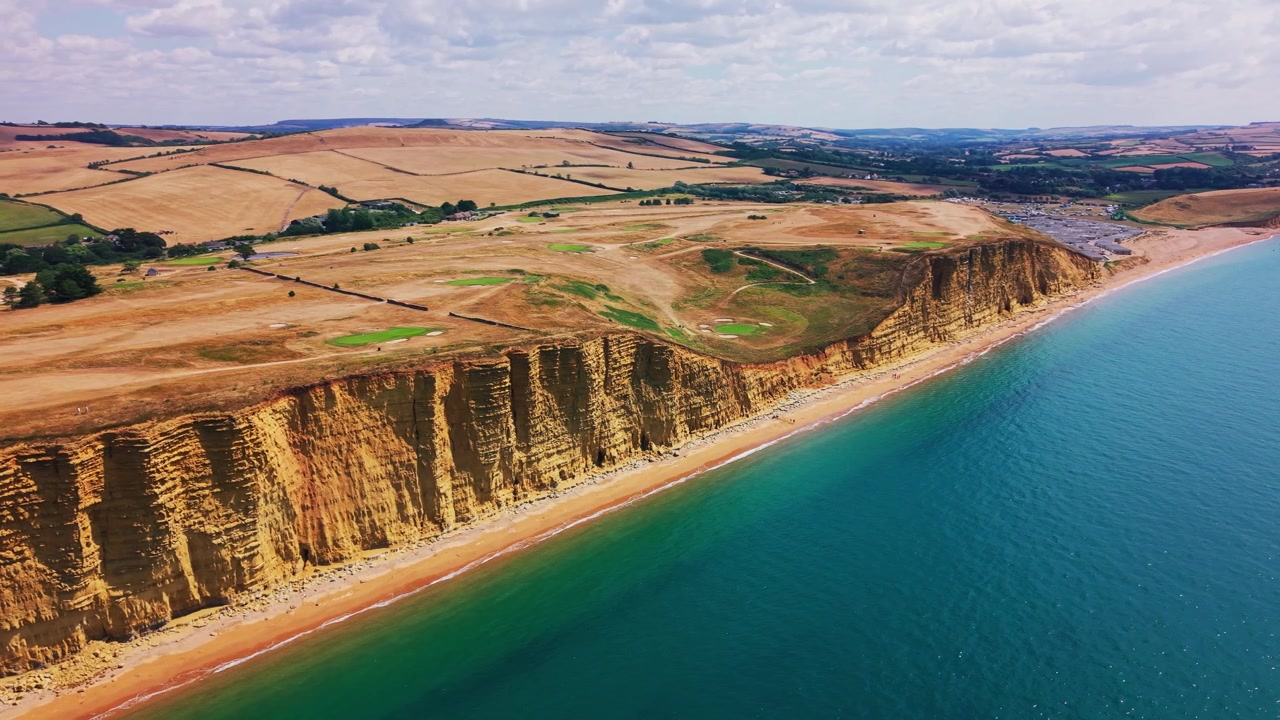 Omfattende luftoptagelser med drone af de gyldne klipper, det turkisblå hav og det bølgende landskab ved West Bay på Dorsets Jurassic Coast, England. Den brede kystudsigt afslører lagdelte klippeformationer.