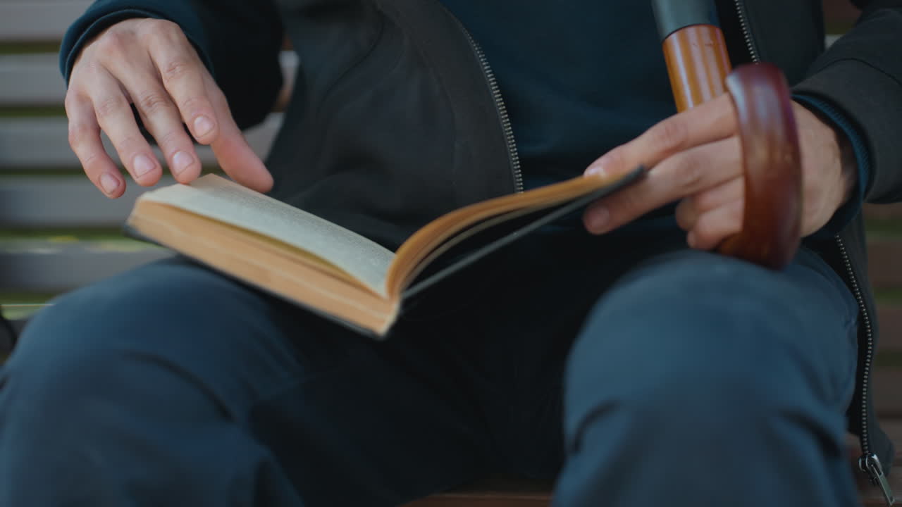 vieil homme lisant sur un banc de parc, gros plan sur ses mains tournant les pages d’un livre à couverture en cuir à côté d’une canne en bois, lumière calme de l’après-midi, solitude méditative, pages texturées et gestes lents